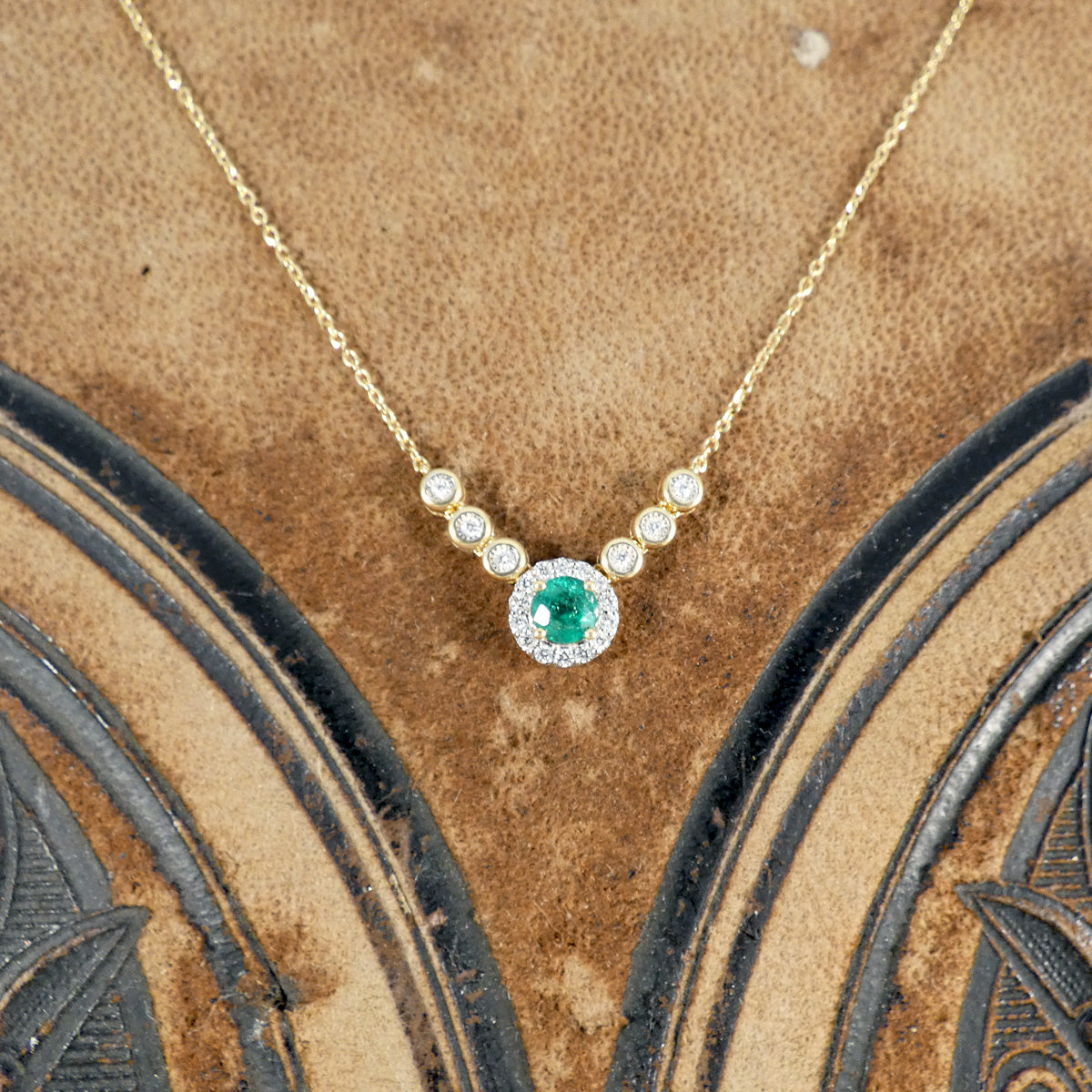 A stunning new necklace crafted in 18ct yellow gold, featuring a vibrant green emerald centre piece surrounded by a halo of sparkling diamonds, complemented by additional bezel-set diamonds in an illusion setting along the chain.