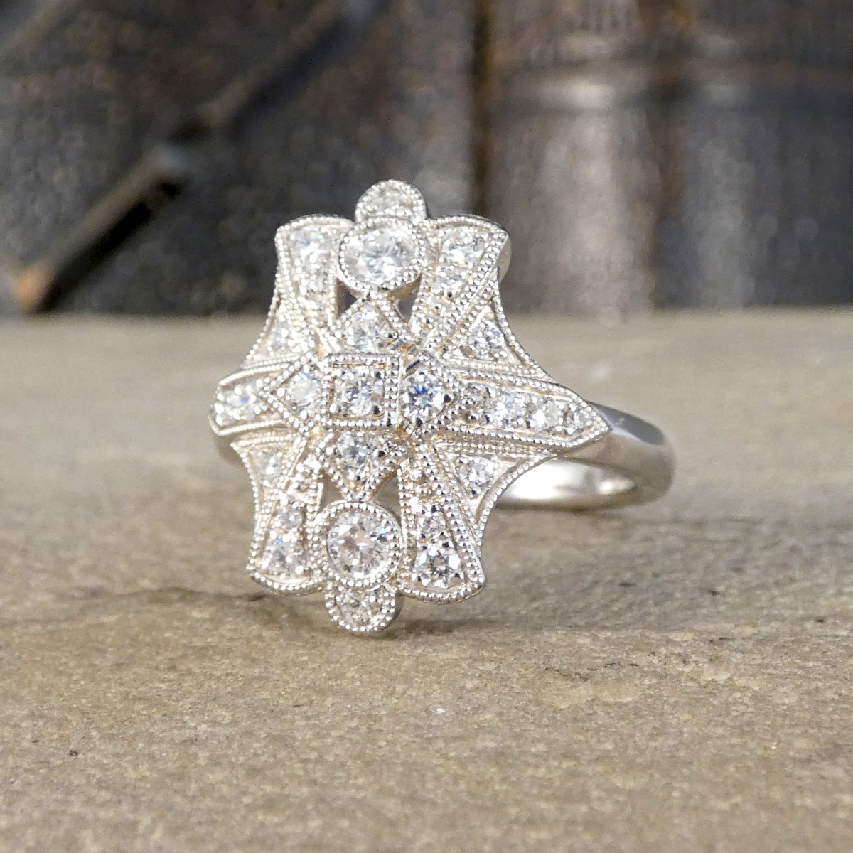 An ornate Art Deco style platinum ring with multiple clear gemstones, resting on a textured stone surface with a blurred dark background.