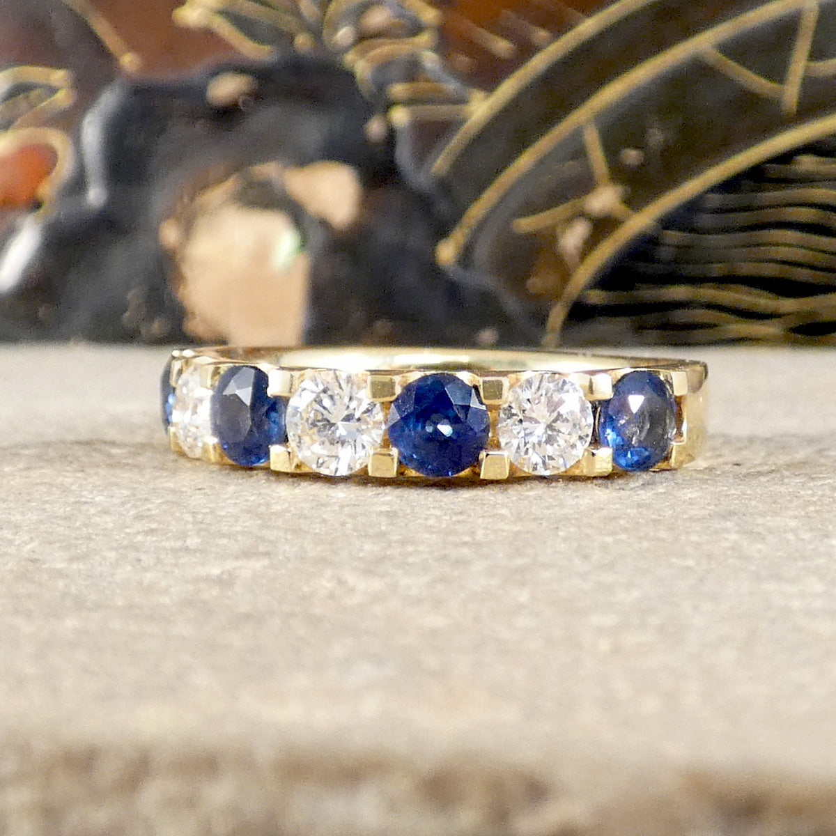 A gold eternity ring featuring alternating round blue sapphires and clear round diamonds, resting on a textured beige surface with a blurred dark background.