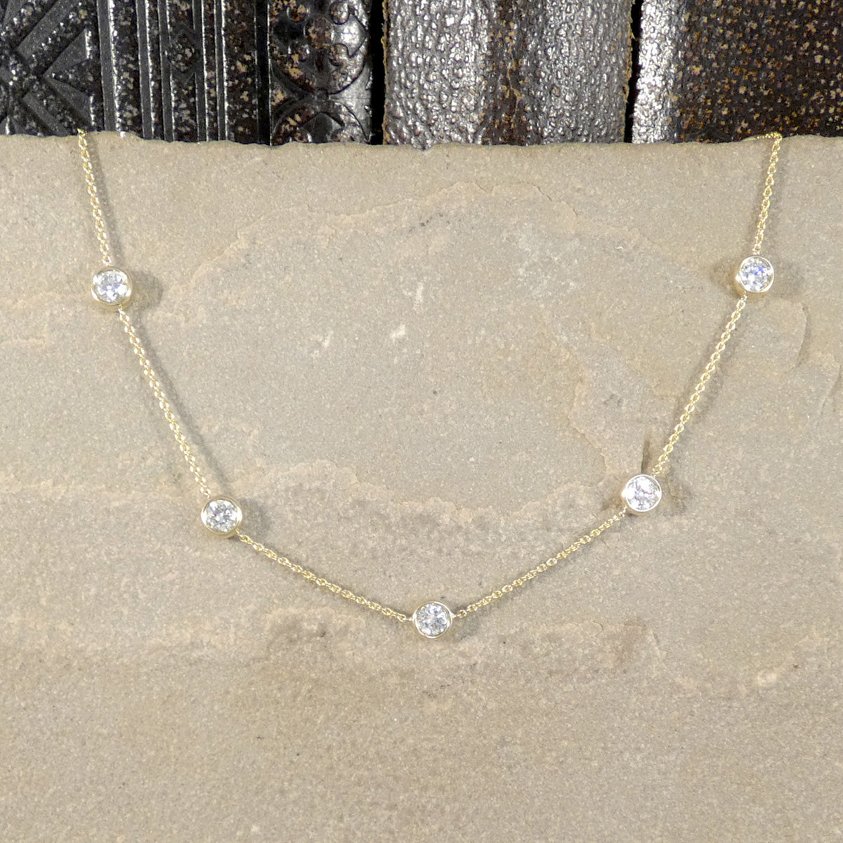 A delicate gold chain necklace with five small, round clear gemstones spaced evenly along the chain, displayed on a textured beige stone surface.