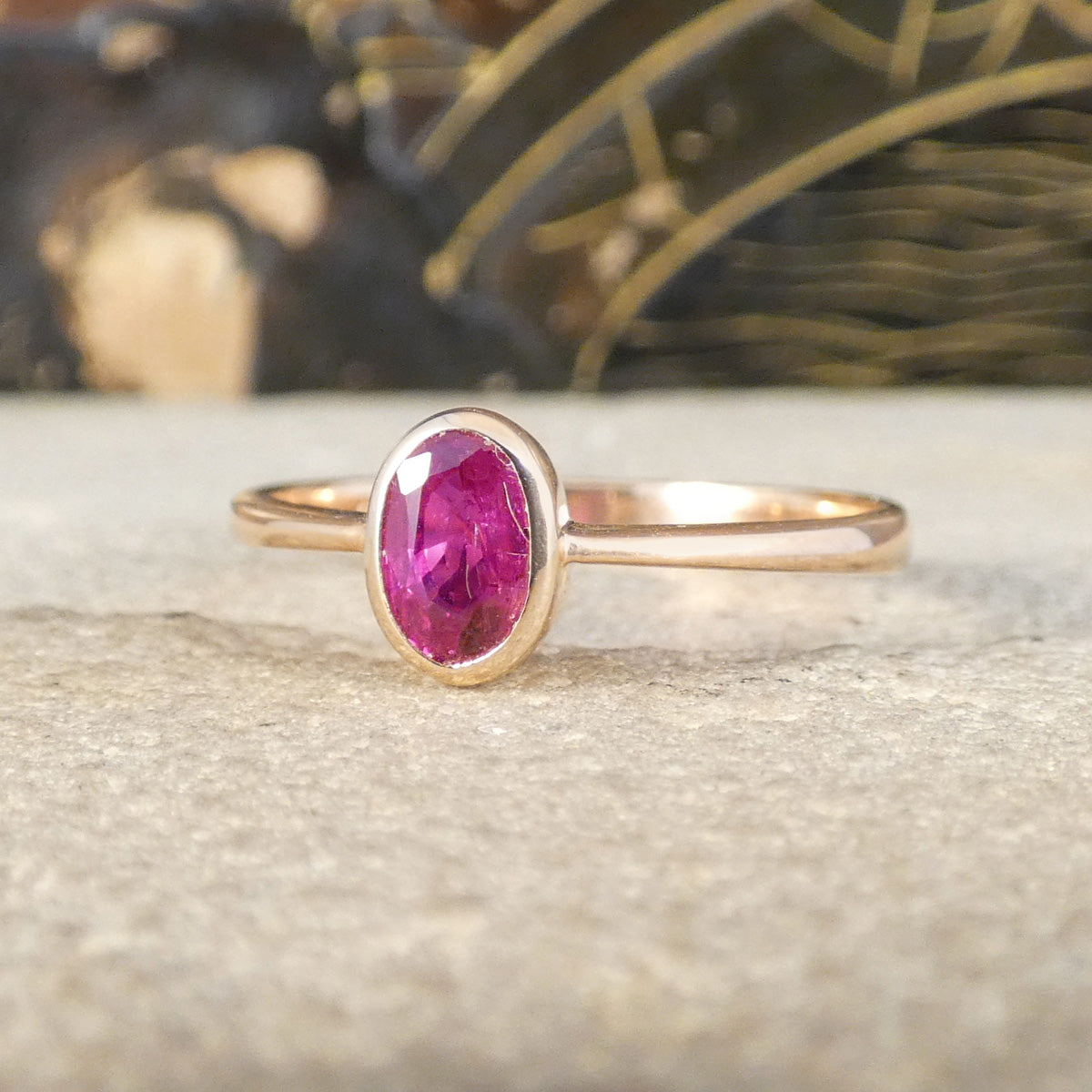 A rose gold ring with an oval-cut pink gemstone, resting on a textured stone surface with a blurred background.