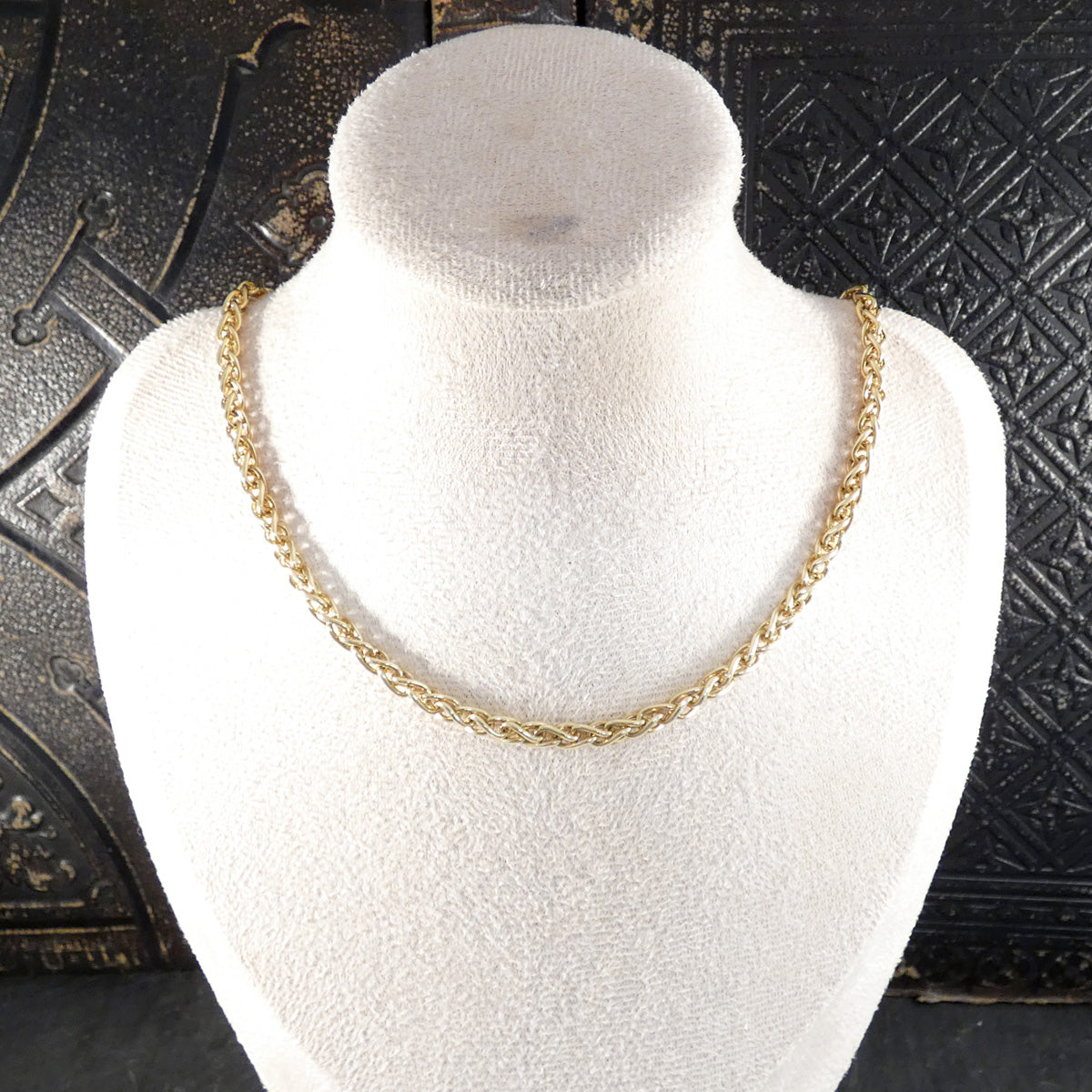 A gold chain necklace displayed on a white fabric-covered jewelry bust against a dark textured background.