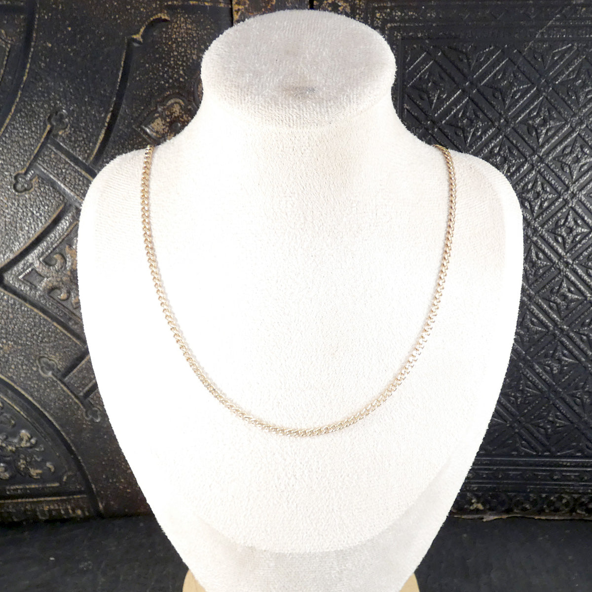 A delicate gold chain necklace displayed on a white fabric jewelry bust against a dark textured background.