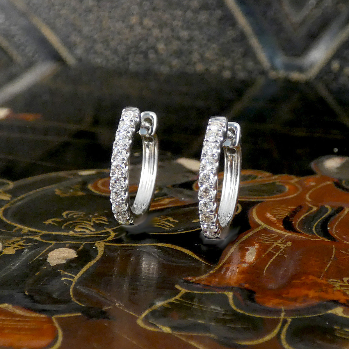 A pair of silver huggie hoop earrings paved with small clear stones, displayed on a glossy, ornate brown and gold patterned surface.