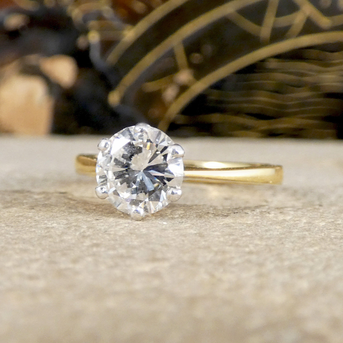 A yellow gold solitaire engagement ring with a round brilliant-cut clear gemstone, resting on a textured beige stone surface with a blurred dark background.
