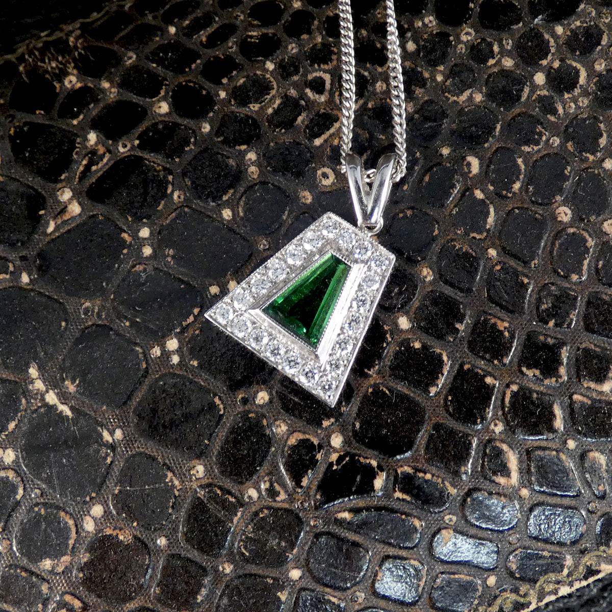 A silver geometric pendant necklace featuring a green malachite triangle in the center, surrounded by a halo of small clear stones, displayed on a textured black reptile skin surface.