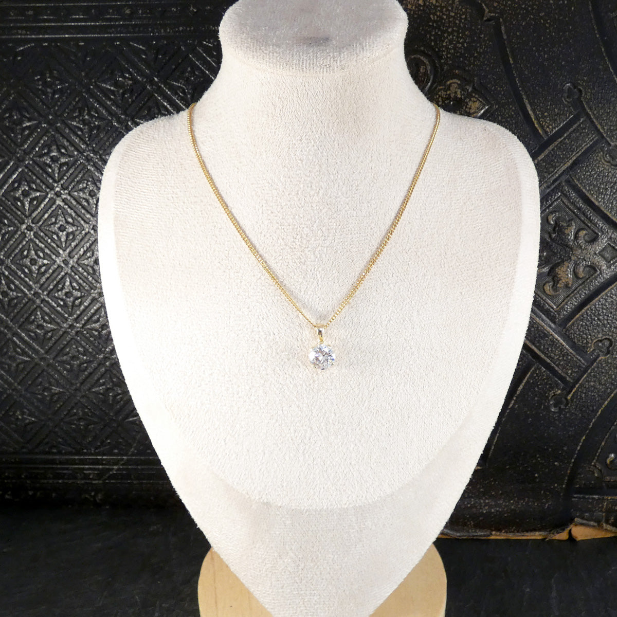 A gold chain necklace with a clear round-cut gemstone diamond pendant, displayed on a white fabric jewelry bust against a dark textured background.