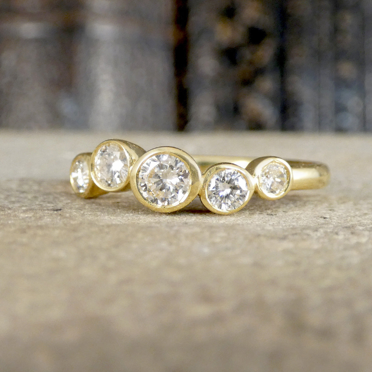 A yellow gold ring featuring five round-cut clear gemstones in bezel settings, arranged in a graduated pattern, resting on a textured stone surface.