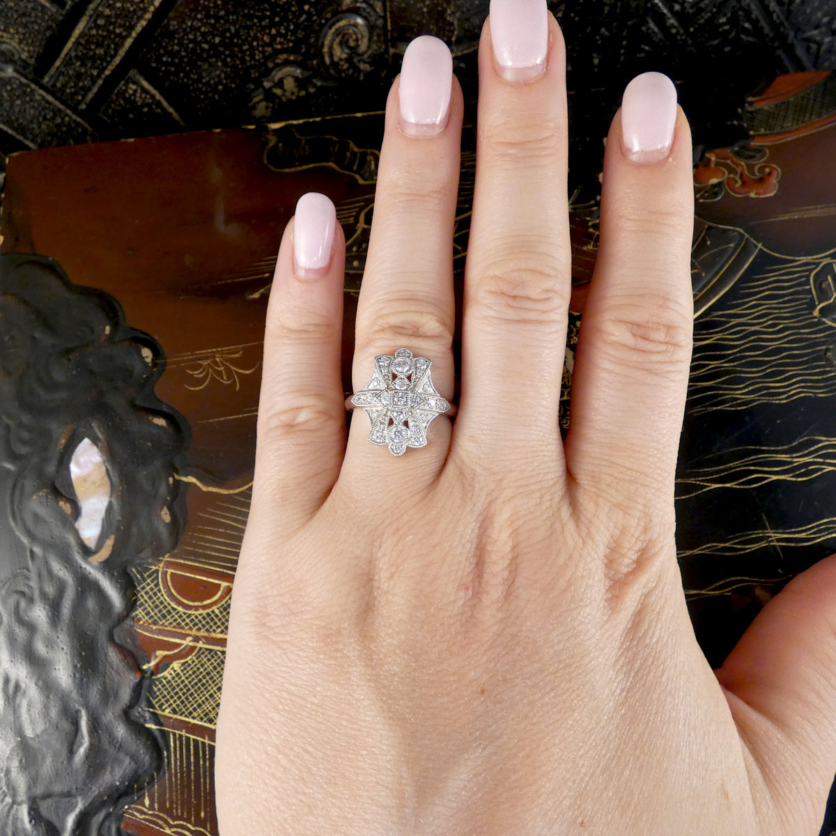 A close-up of a hand with pale pink manicured nails wearing a silver-toned ring with a floral cluster of small clear stones and tiny red gem accents, set against a dark patterned background.