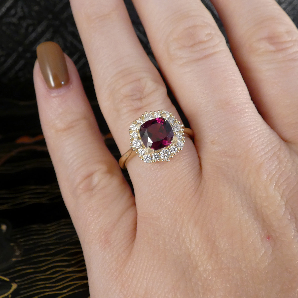 A close-up of a hand wearing a gold ring with a cushion-cut dark red gemstone surrounded by a halo of small clear stones.