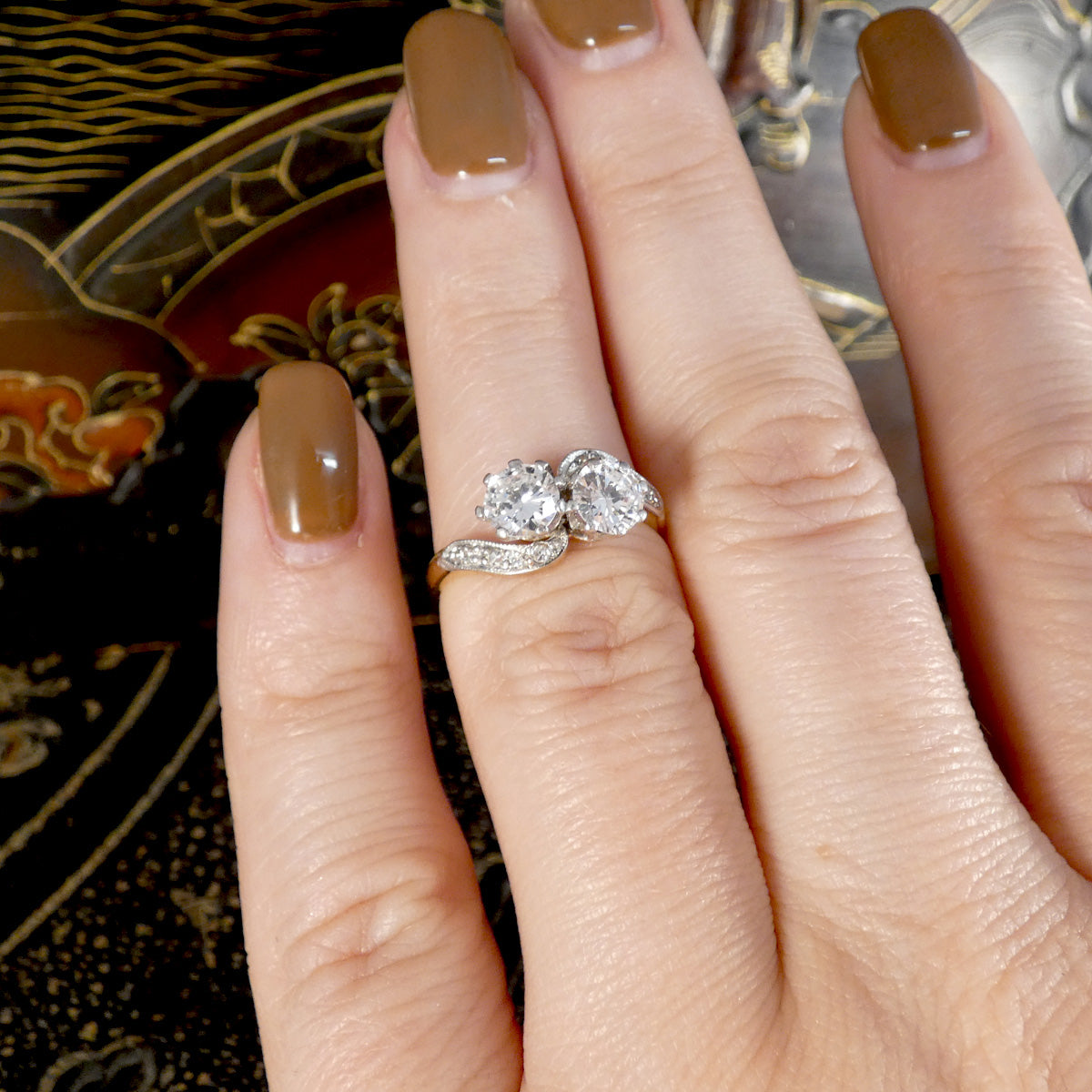 A close-up of a hand wearing a silver ring with two round clear gemstones and a pavé band, set against a dark patterned background.