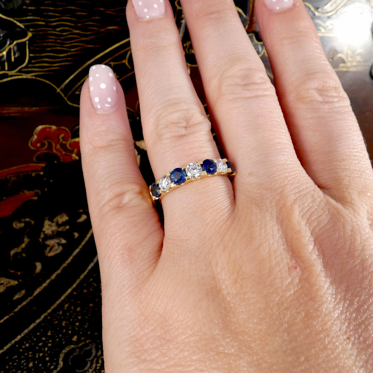 A close-up of a hand wearing a yellow gold ring set with round-cut blue sapphires and clear stones, photographed against a dark patterned background.