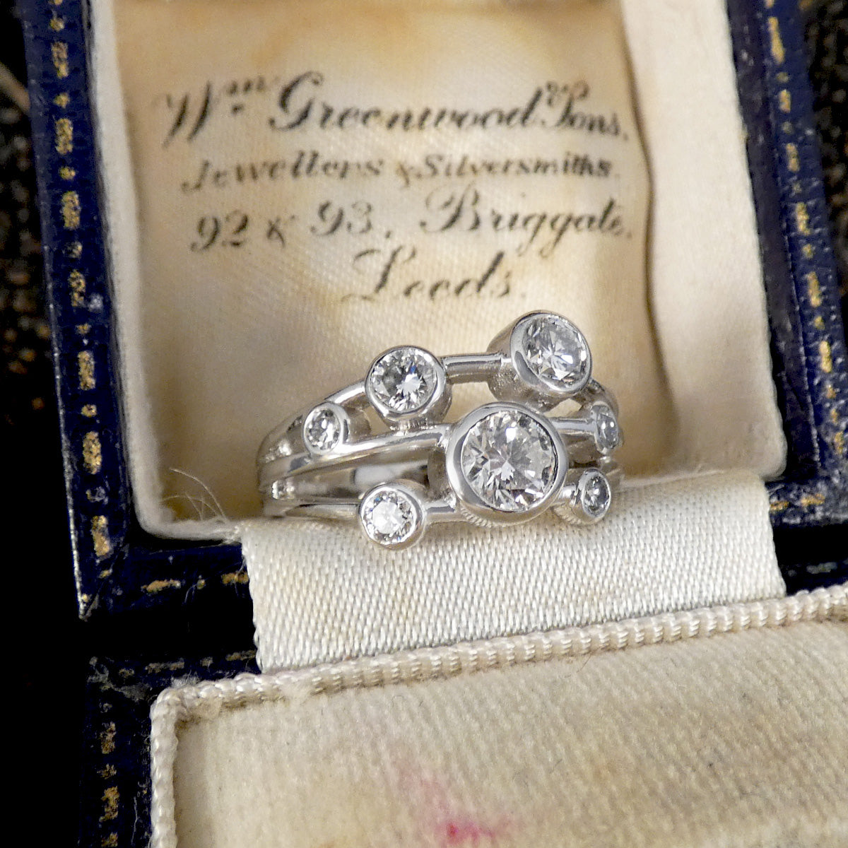A silver-toned multi-stone ring with round diamond gemstones, displayed inside an open vintage jewellery box with cream lining and a dark blue velvet exterior. 