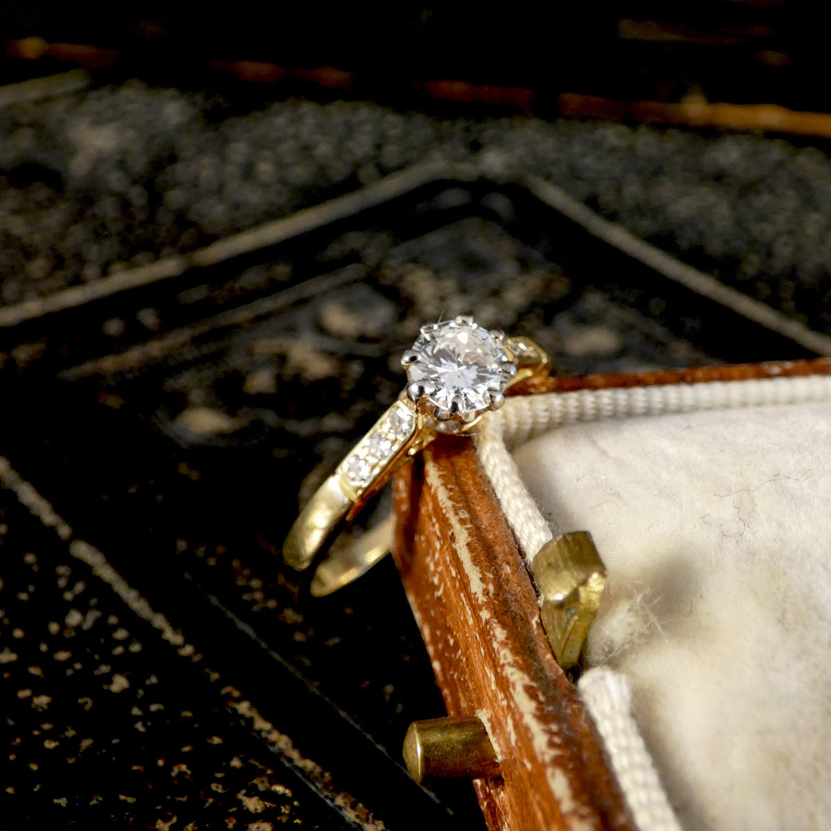 A yellow gold solitaire engagement ring with a round brilliant-cut diamond and a pavé band, resting on a vintage wooden box lid.