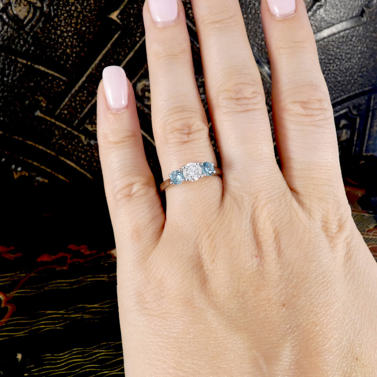 A close-up of a hand wearing a silver three-stone ring with light blue gemstones, set against a dark textured background.