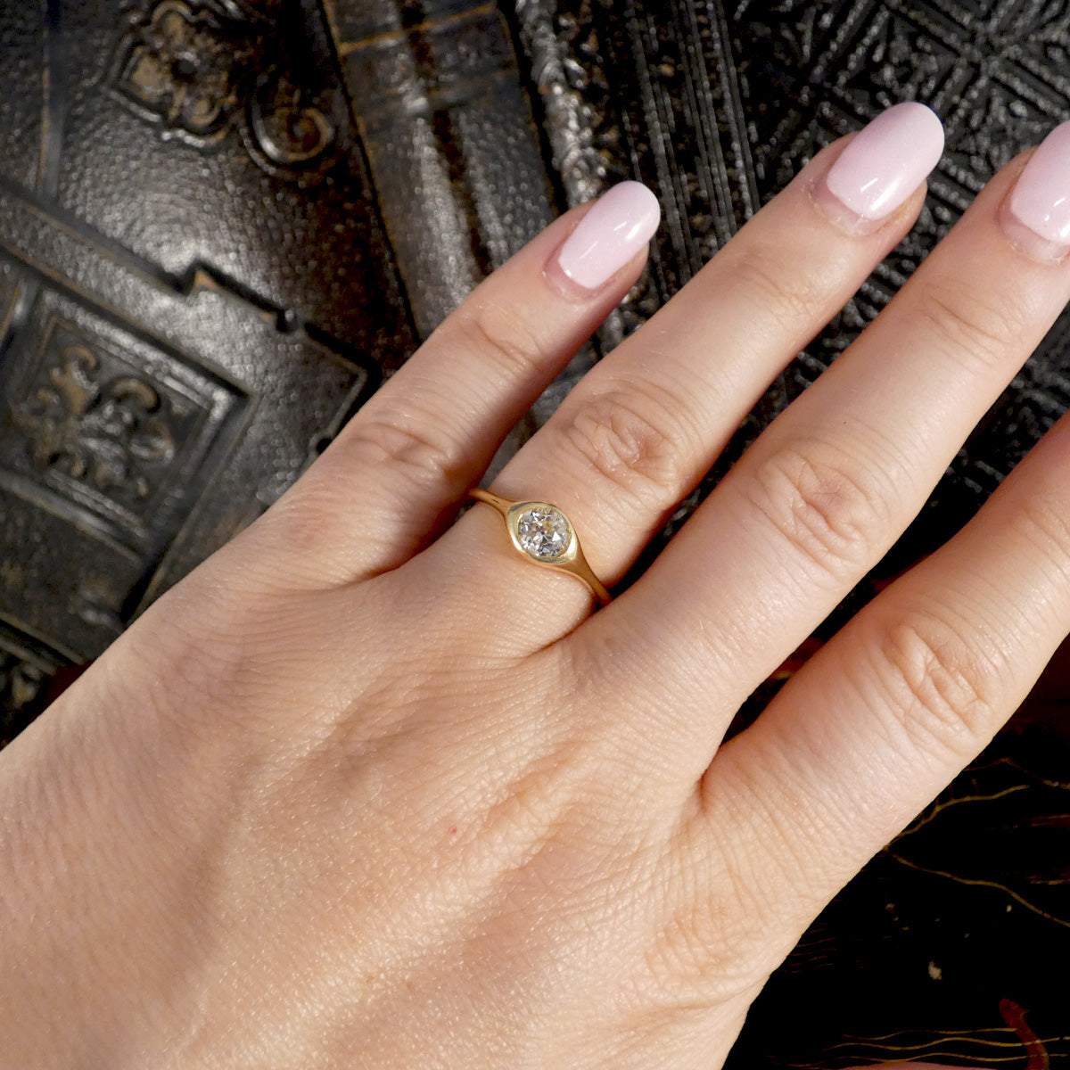 A close-up of a hand wearing a delicate yellow gold ring with a round clear gemstone, set against a dark textured background.