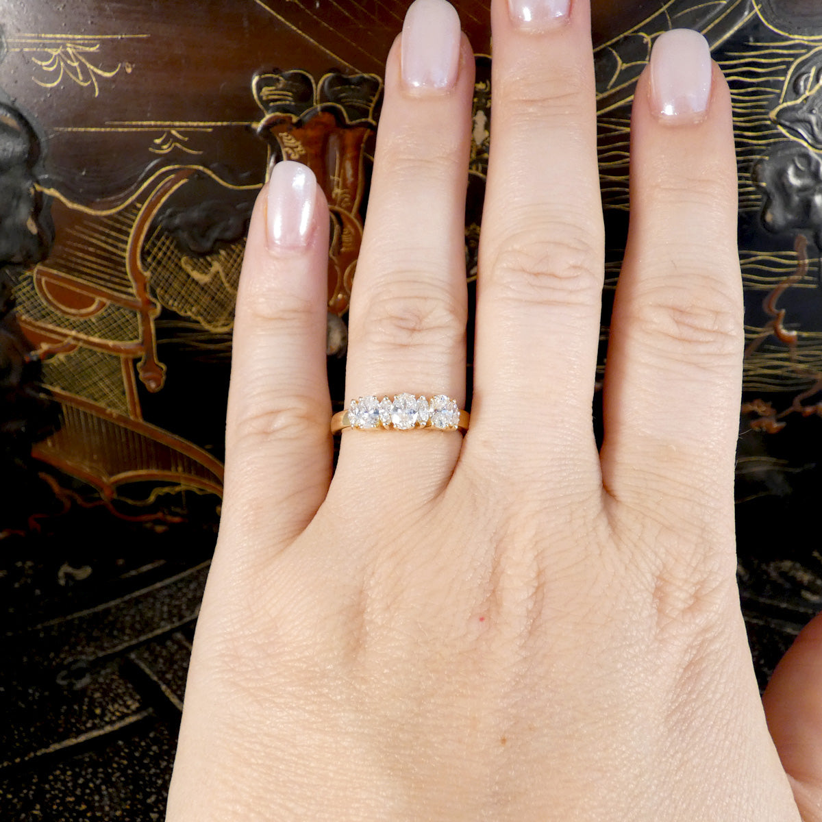 A close-up of a hand wearing a gold three-stone diamond ring, with manicured fingernails and a dark patterned background.