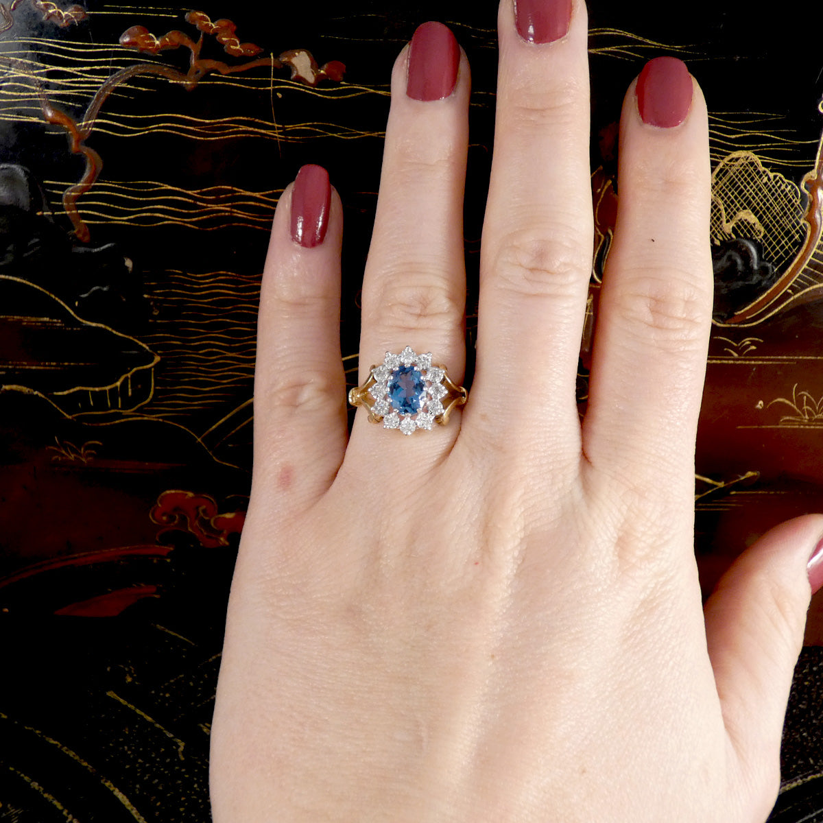 Hand wearing a ring with a blue gemstone against a decorative background