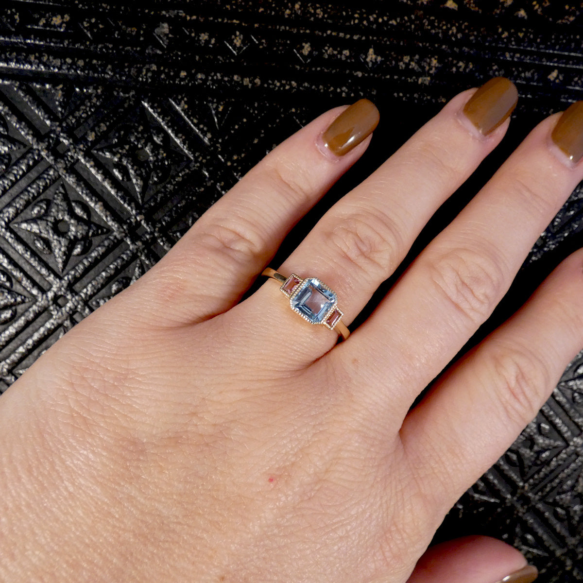 A close-up of a hand wearing a gold ring with a square-cut light blue gemstone, set against a textured black background.
