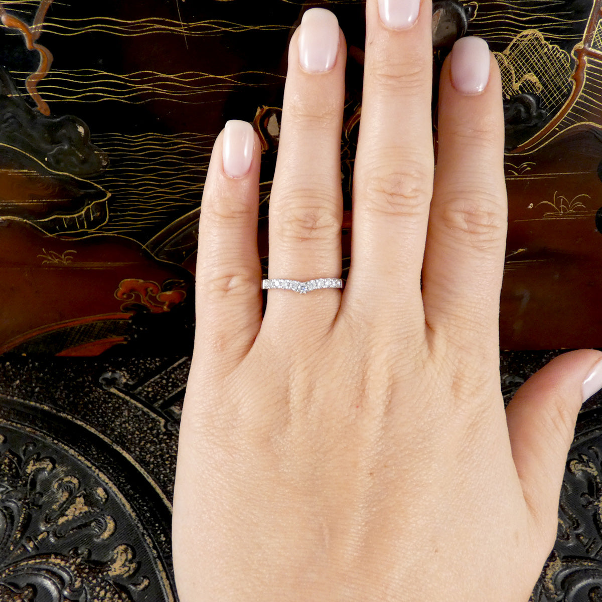 A close-up of a hand with pale pink manicured nails wearing a thin platinum diamond set wave band ring set with small clear stones, resting against an ornate dark brown background.