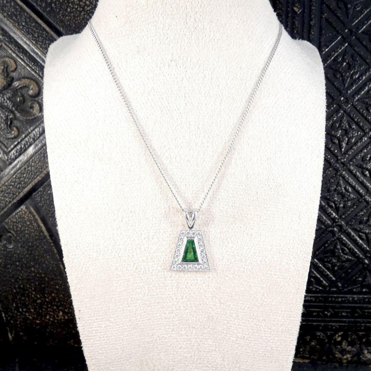 A silver-toned pendant necklace displayed on a white jewelry bust. The pendant features a triangular green gemstone surrounded by small clear stones in a silver setting, suspended from a delicate silver chain.
