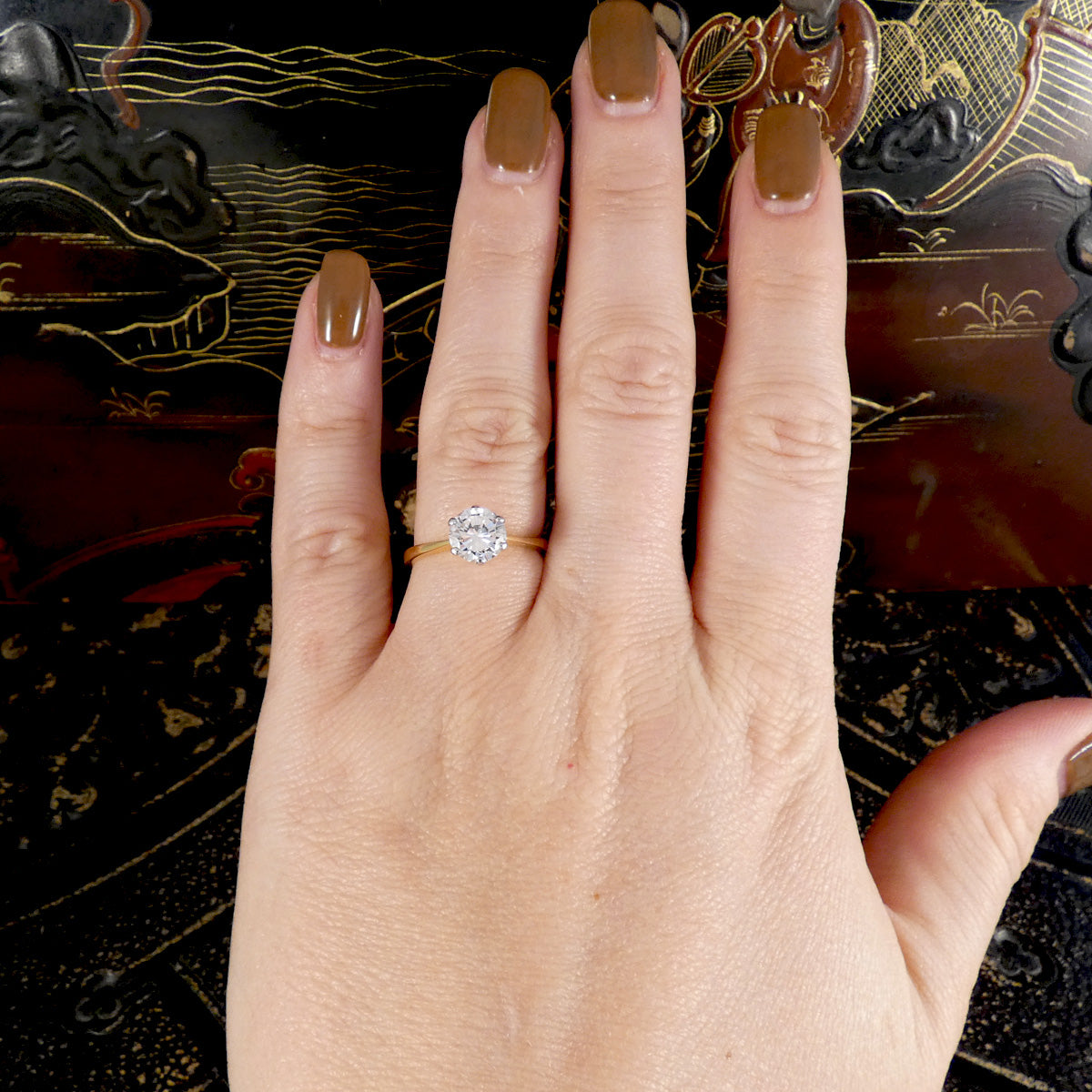 A close-up of a hand with brown manicured nails wearing a gold ring with a round clear diamond, set against a dark patterned background.