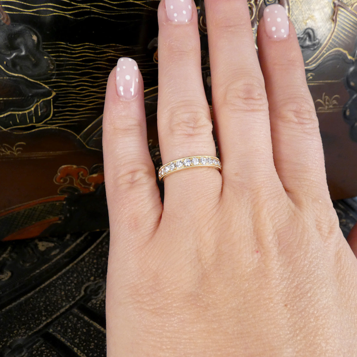 A close-up of a hand wearing a yellow gold eternity ring set with small clear stones. The hand has pale pink manicured nails, and the background shows a dark, ornate patterned surface.