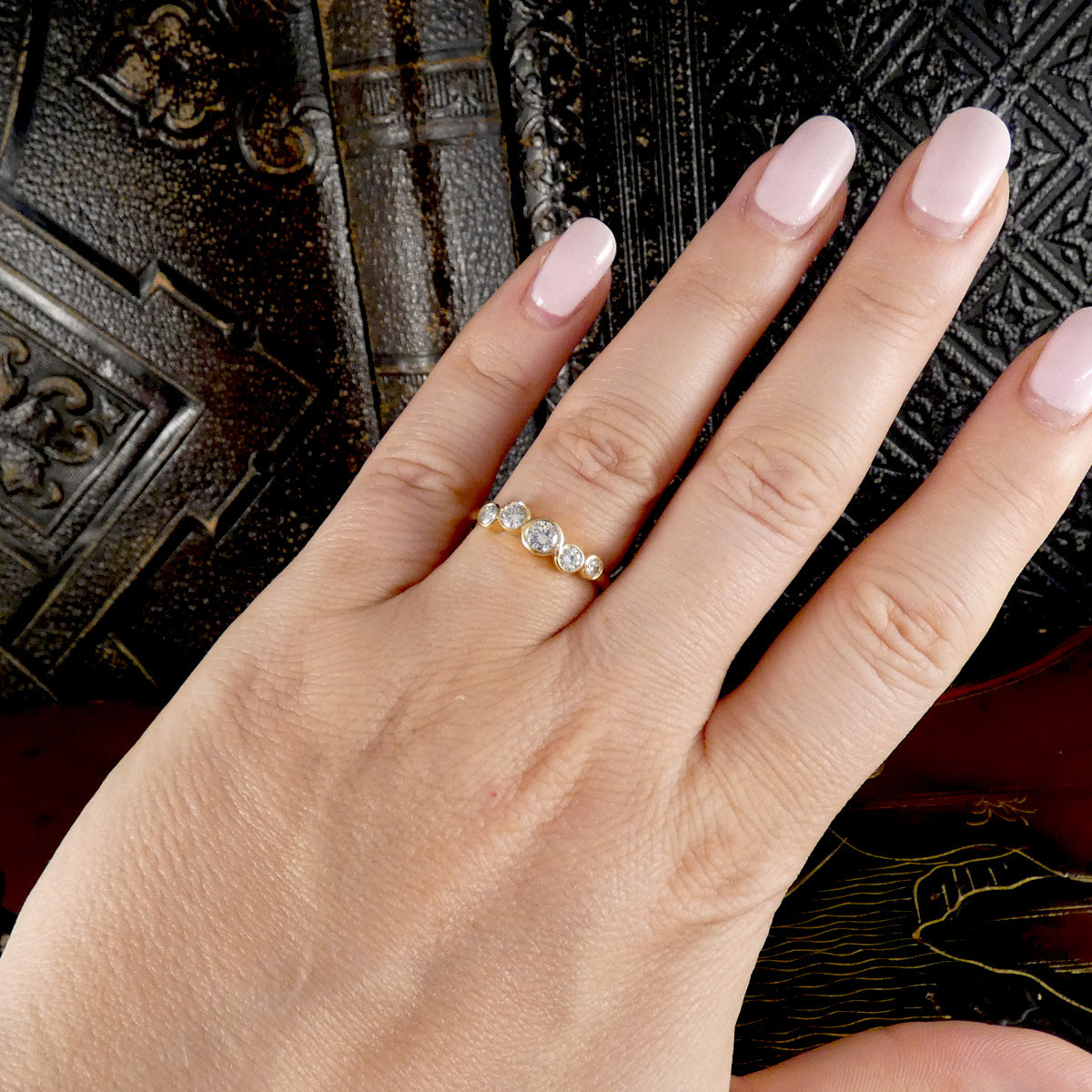 A close-up of a hand with pale pink manicured nails wearing a delicate gold ring set with three clear round gemstones, posed against a dark textured book background.