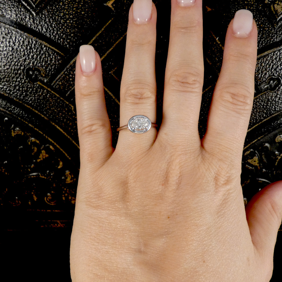 A close-up of a hand wearing a silver-toned oval ring with a clear round-cut center stone, set against a dark textured background.