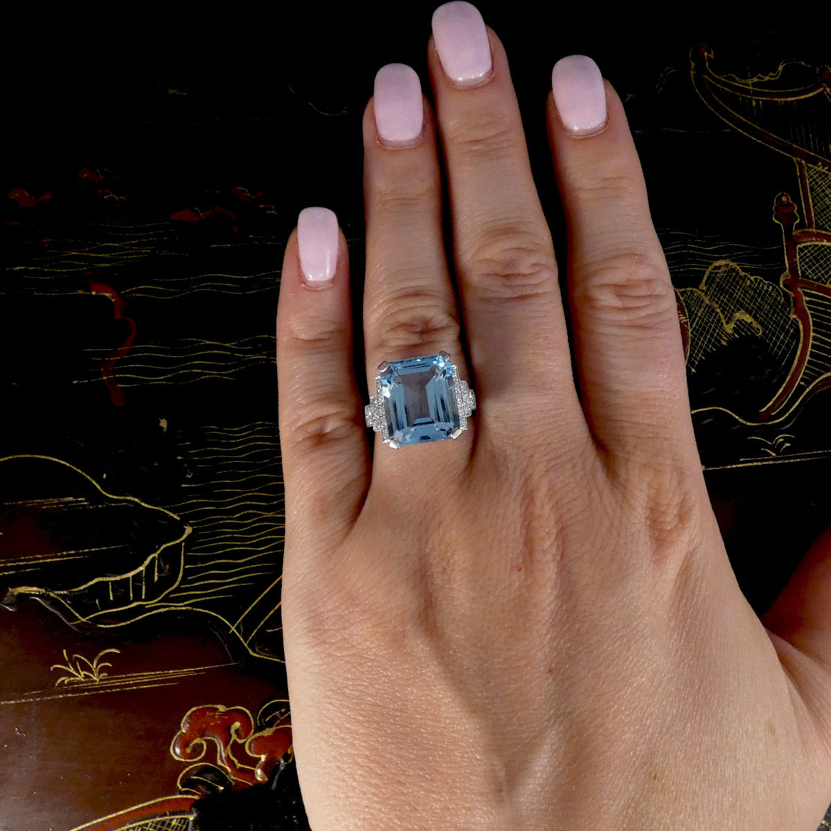 A hand with light pink manicured nails wearing a silver ring featuring a large rectangular light blue gemstone with small clear stones on either side, set against a dark patterned background.