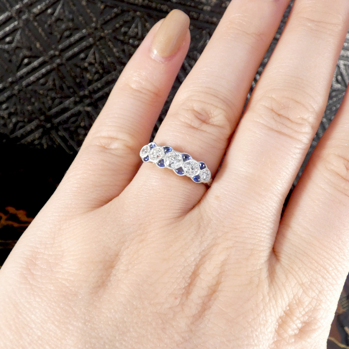 Platinum ring featuring a row of old-cut diamonds interspersed with blue sapphire accents, worn on the finger to showcase its elegant Art Deco-inspired design and intricate craftsmanship.