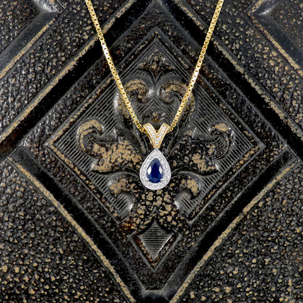 A gold chain necklace with a teardrop-shaped pendant featuring a deep blue center stone surrounded by small clear stones, displayed on a textured dark metallic background.