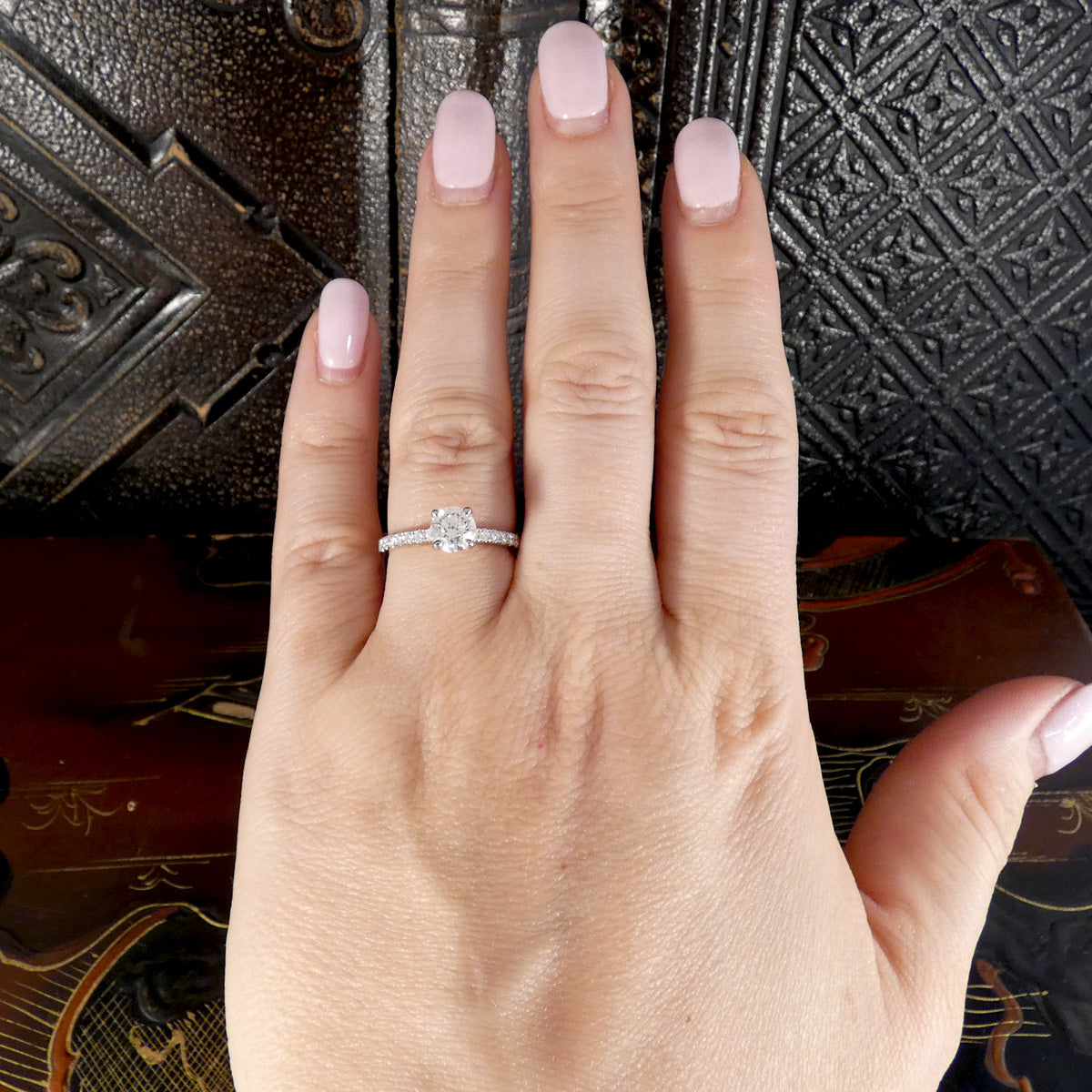 A close-up of a hand wearing a silver-toned diamond ring with a round center stone and smaller side stones, set against a dark textured background.
