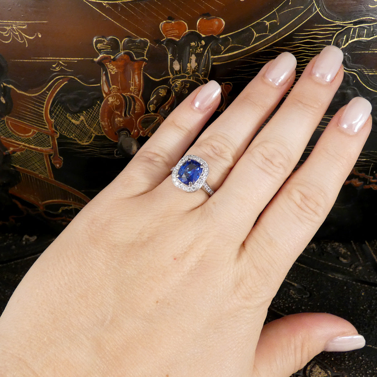 A close-up of a hand wearing a silver ring with a cushion-cut blue sapphire and a halo of small clear stones, set against a dark patterned background.