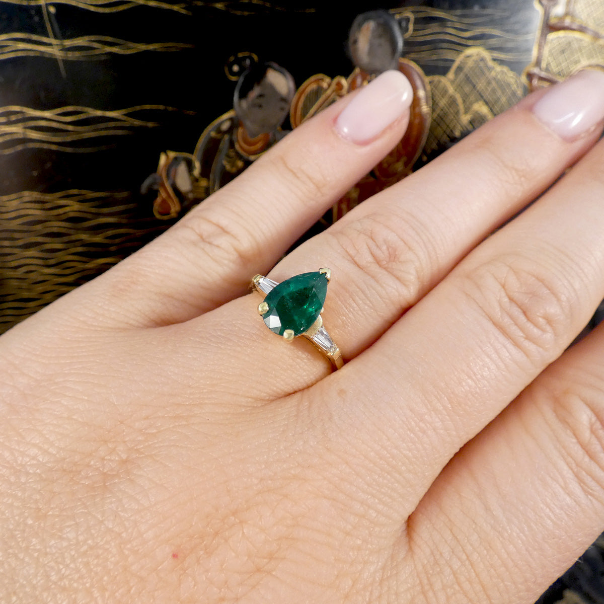 Hand wearing a ring with a green emerald and diamond gemstone against a decorative background