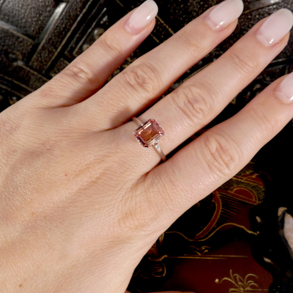 A close-up of a hand wearing a ring with an emerald-cut pink gemstone on a thin gold band, set against a dark patterned background.
