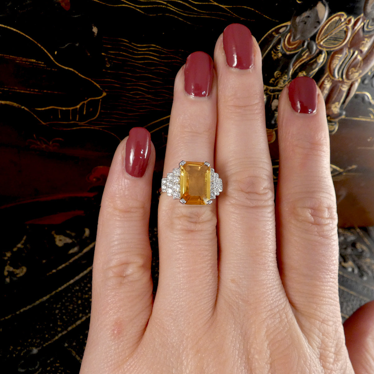 Hand wearing a ring with a yellow sapphire and diamond art deco style setting against an ornate background