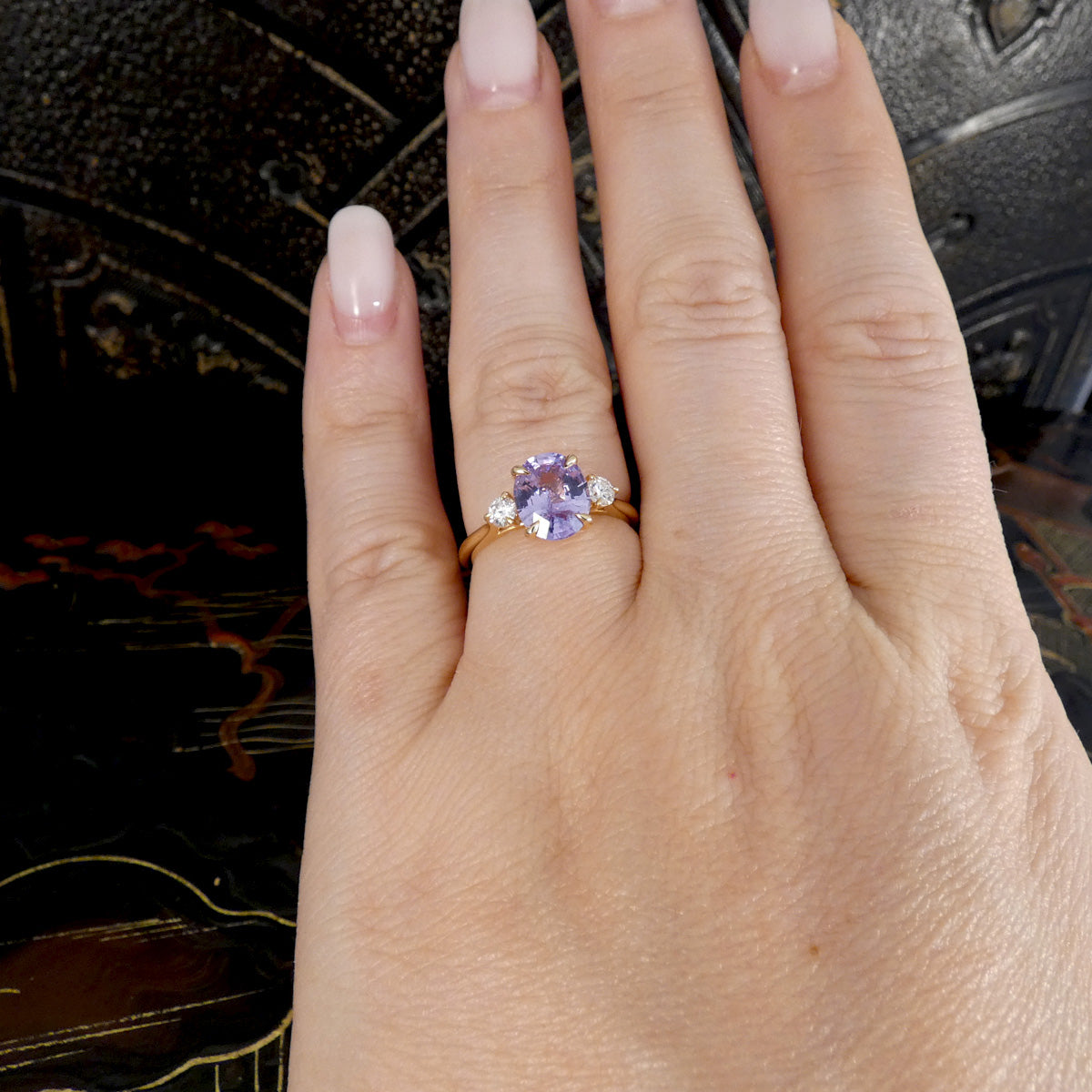 A gold ring with a round lavender-purple center stone and two small clear side stones, shown worn on a person's ring finger. The hand has a pale pink manicure, and the background is a dark textured surface.