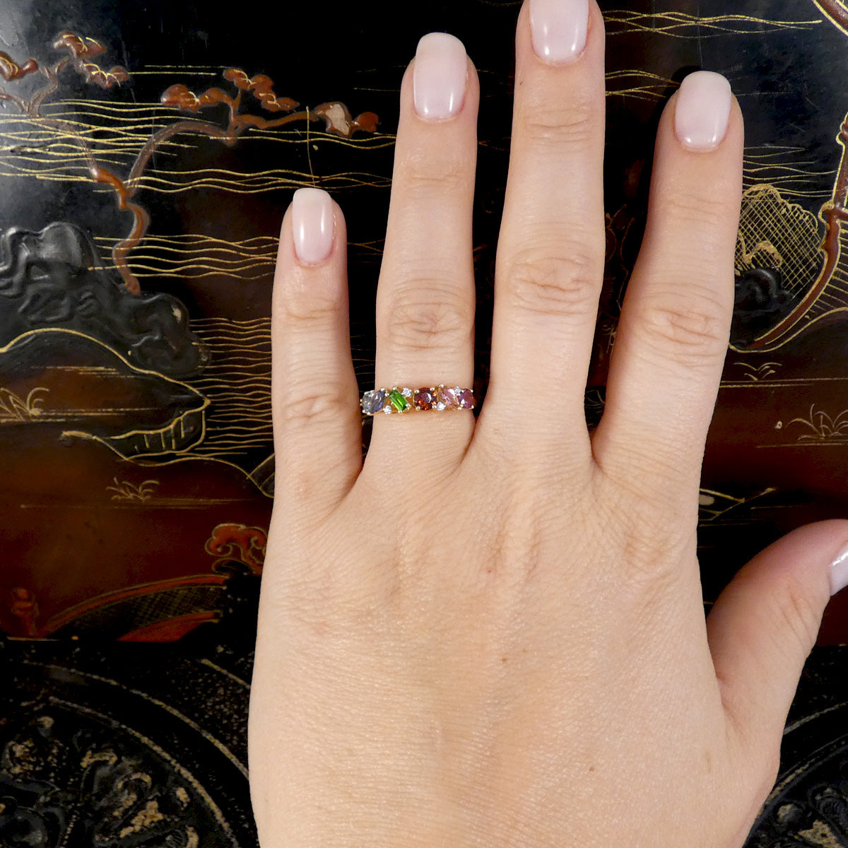Hand wearing a ring with multicolored gemstones against an ornate background