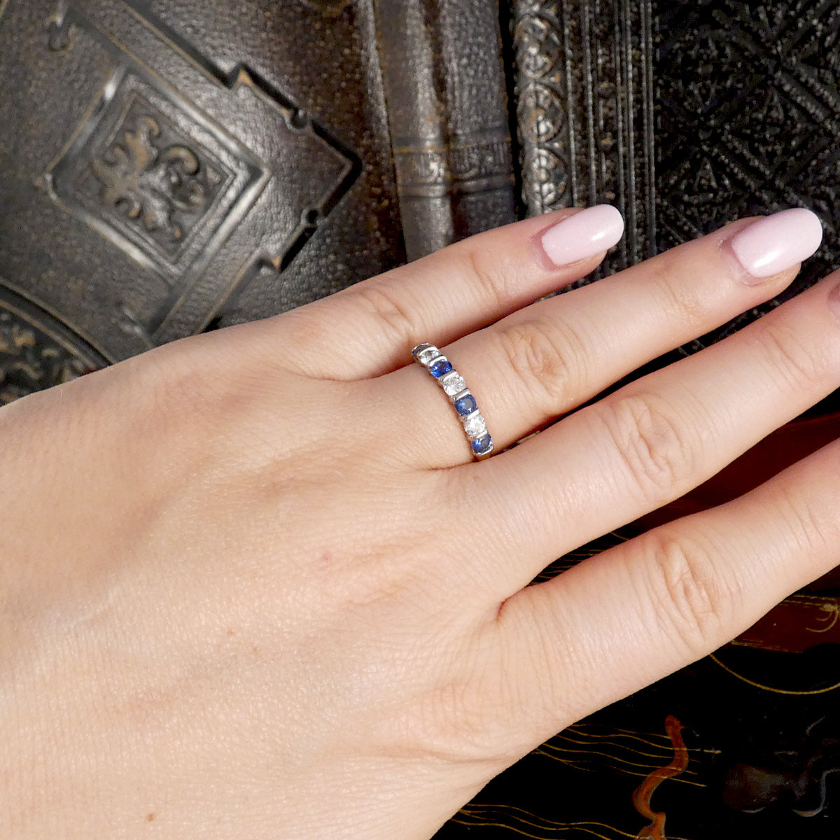 A close-up of a hand wearing a silver band ring with alternating round-cut blue sapphires and clear stones, set against a dark textured background.