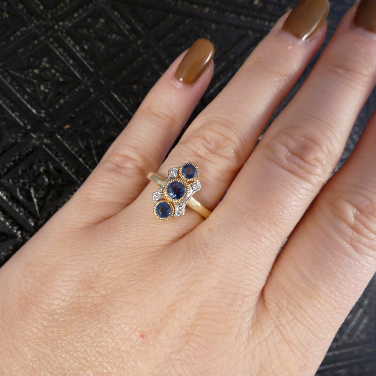 A close-up of a hand wearing a gold ring with three oval blue sapphires and small clear accent stones, set against a dark textured background.