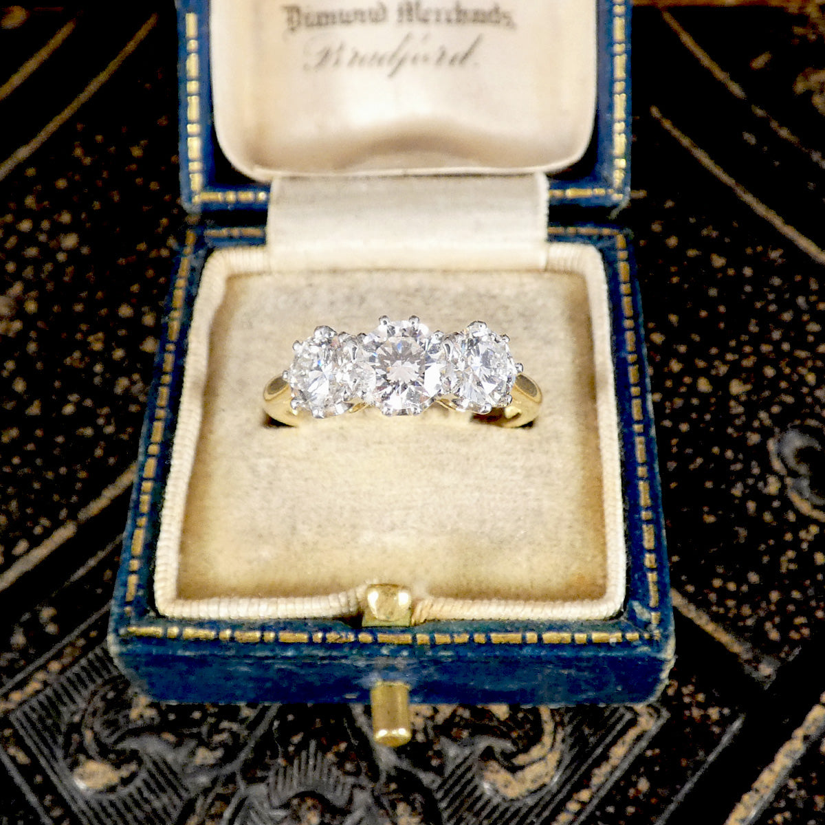 A yellow gold three-stone diamond ring displayed in an open vintage-style blue velvet ring box with a cream velvet interior, set against a dark patterned background.