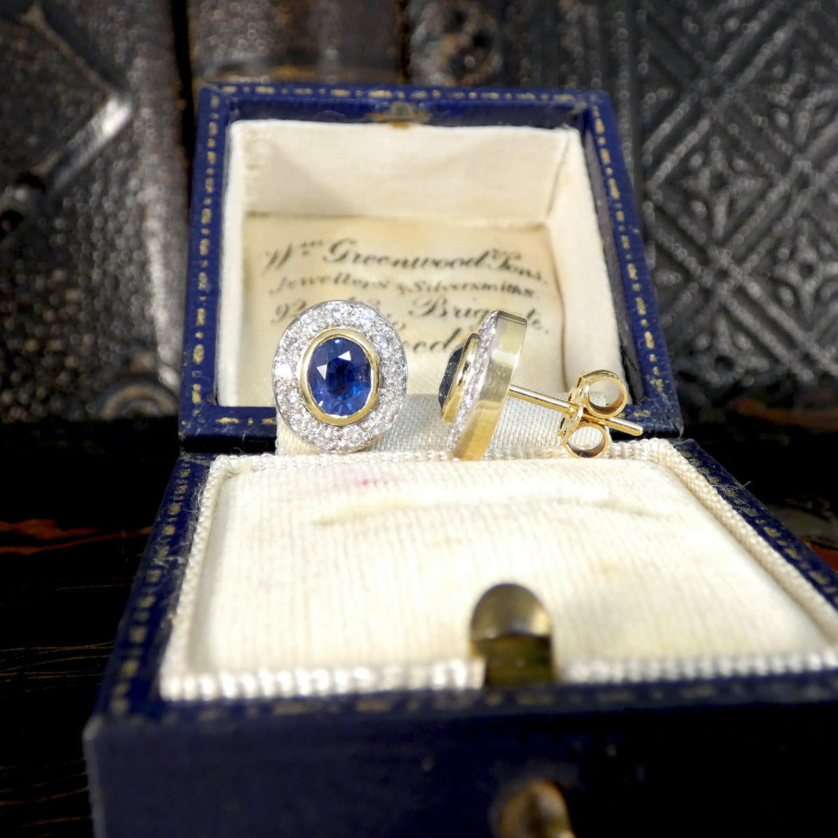 A beautifully bright pair of sapphire and diamond stud earrings in 18ct gold with a bezel setting. Displayed in a vintage box, one from the front and one from the side.