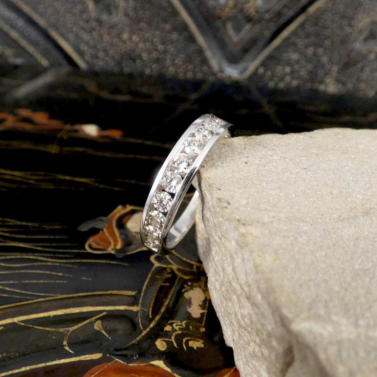 A silver band ring with a channel of clear pavé stones, resting on a light gray stone against a dark, patterned background.