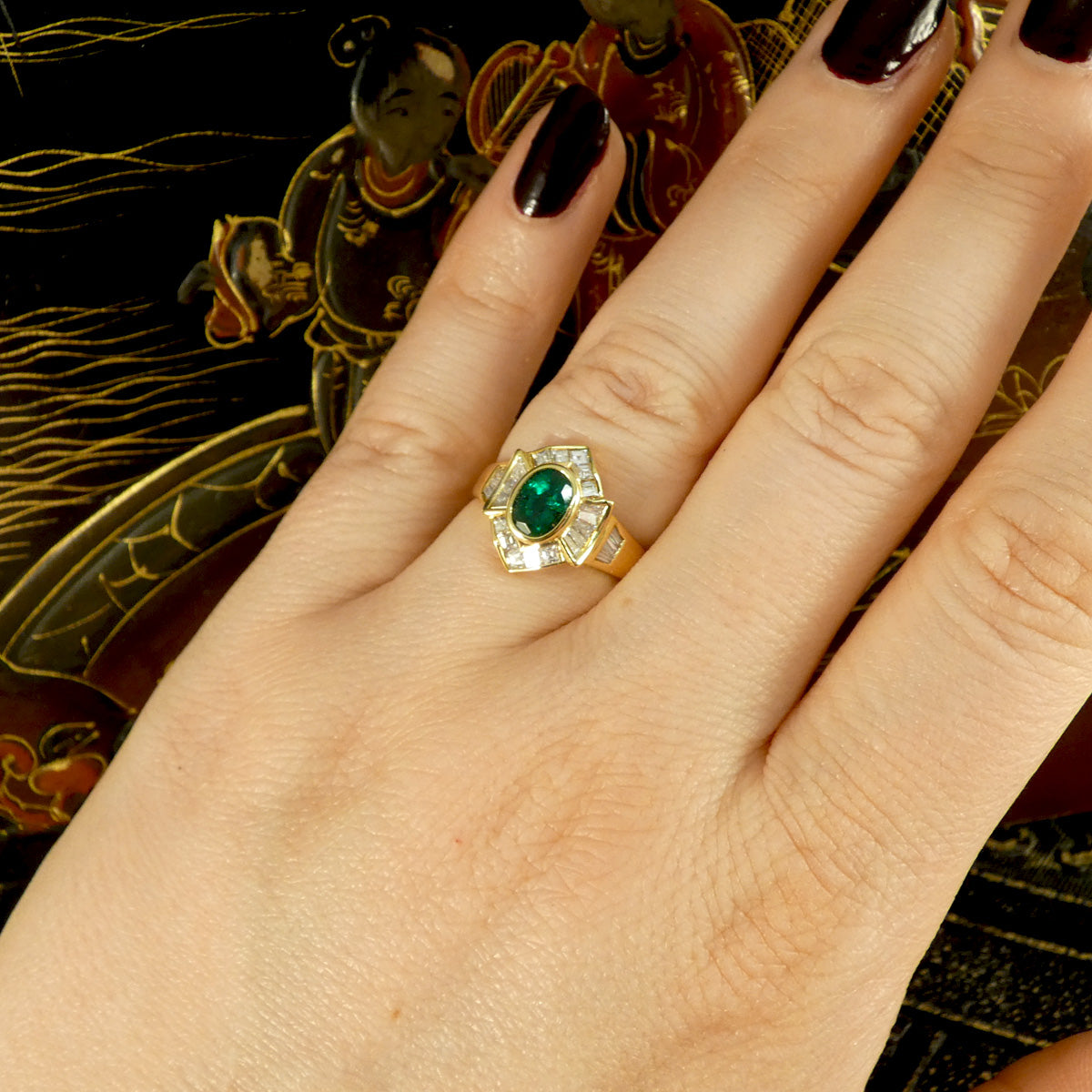 Elegant emerald and tapered diamond cluster ring set in yellow metal, testing as 18ct yellow gold, featuring a vivid oval emerald centre stone surrounded by shimmering tapered baguette diamonds. Displayed on hand for size and ratio guide.