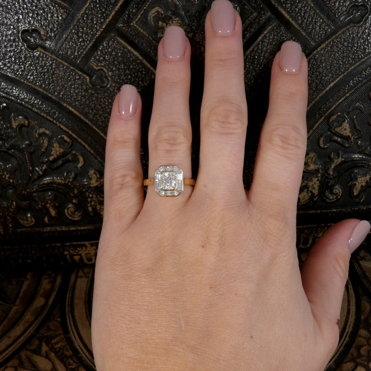 A close-up of a hand wearing a yellow gold ring with a cushion-cut diamond and a halo of smaller round diamonds, set against a dark ornate background.