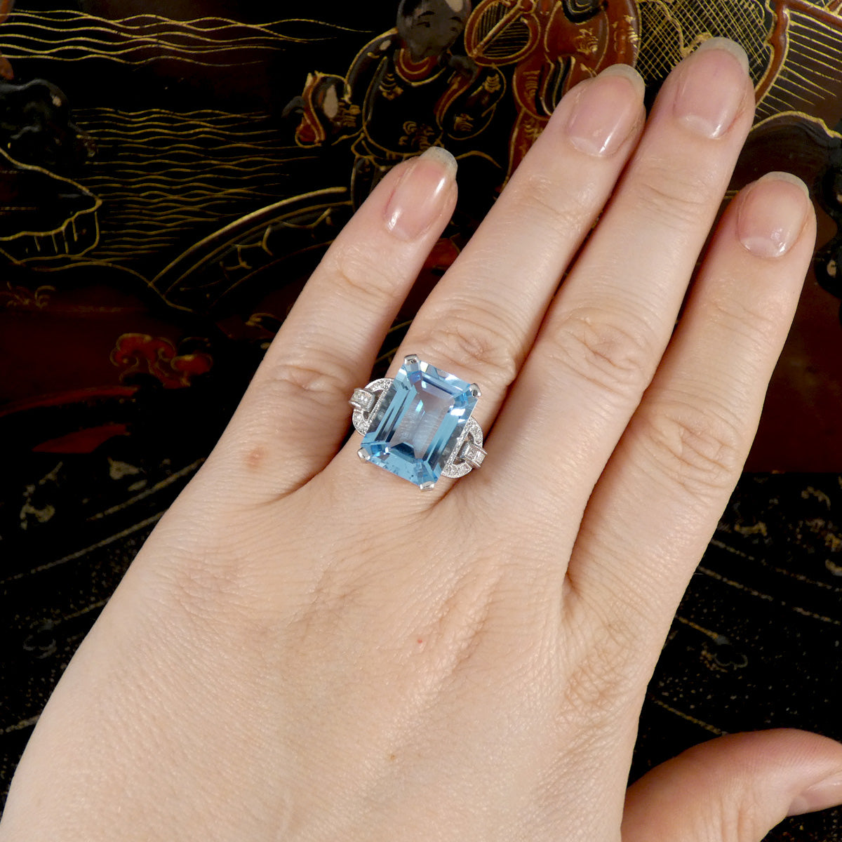 Hand wearing a ring with a blue gemstone against an ornate background