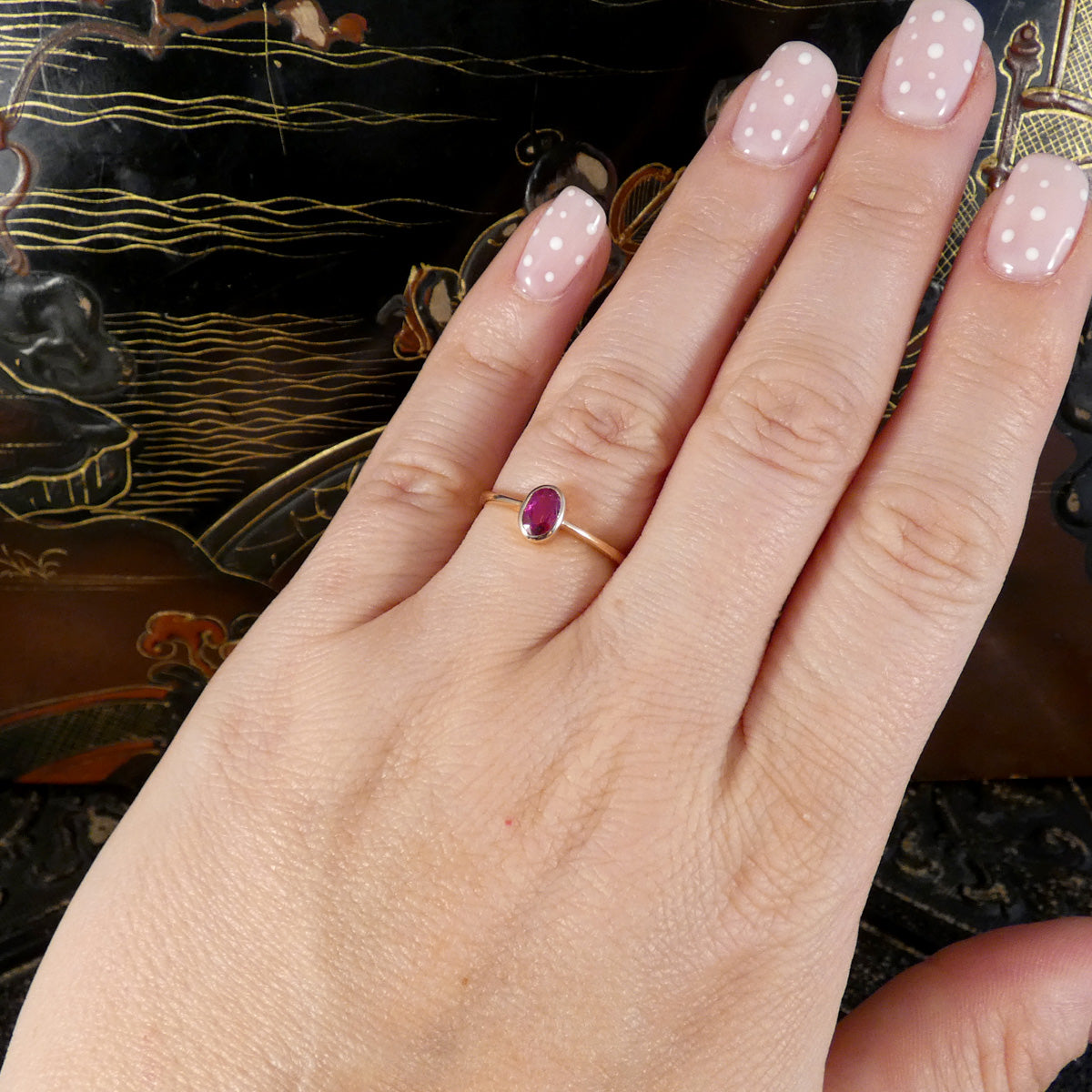 A gold ring with a small oval pinkish-red ruby gemstone worn on a hand with pale pink polka-dot nail polish, set against a dark patterned background.