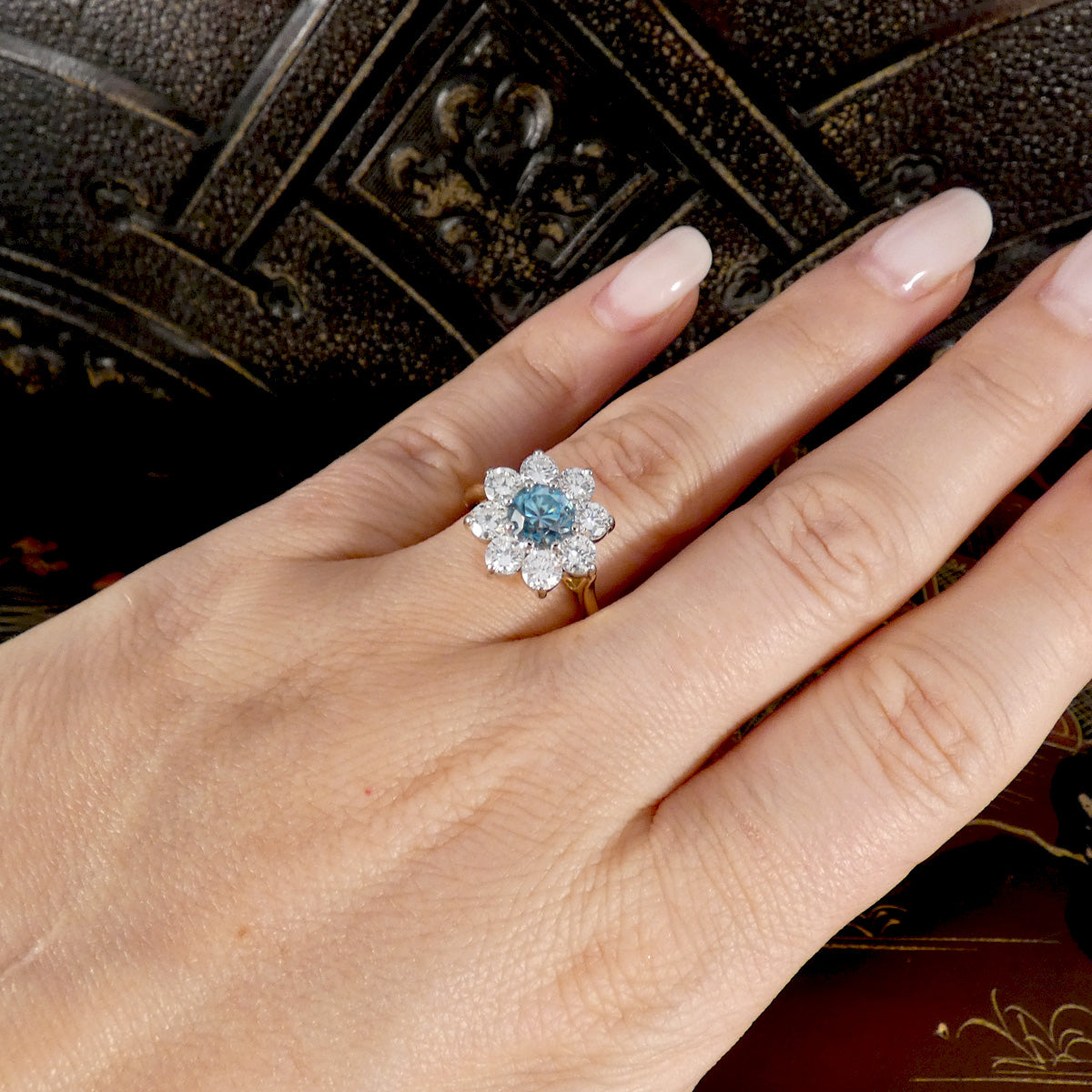 A close-up of a hand wearing a flower-shaped ring with a round blue zircon center stone surrounded by small clear stones, set against a dark textured background.