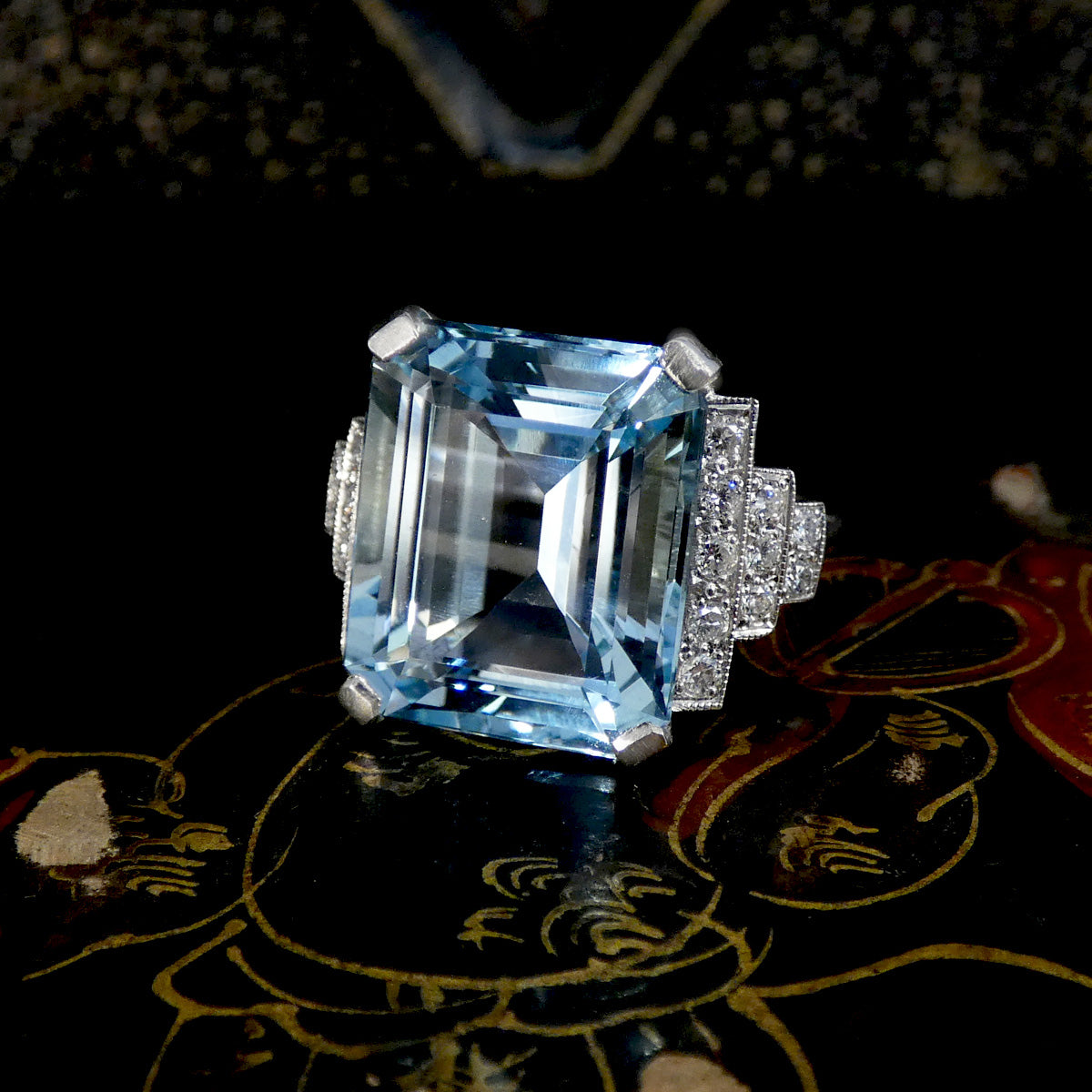 An emerald-cut light blue aquamarine ring with a silver-toned setting and small clear accent stones, displayed on a dark patterned surface.